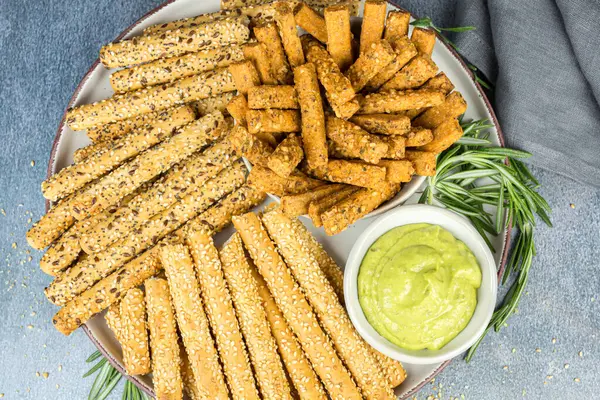 Ekmek çubukları, bitki sosu ve biberiye yaprakları seramik tabakta, kumaş peçete gri arka planda.