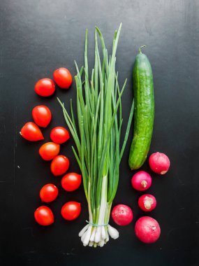 Mevsimlik diyet ve beslenme, sağlıklı yeme konsepti. Bir demet taze kırmızı kiraz domatesleri, yeşil pırasa, turp ve salatalık siyah masa zemininde, sebze manzarası masanın üstünde..