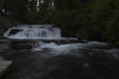 Kayaların üzerinden akan dere ve arka planda şelale. Yemyeşil yeşillikler içinde.