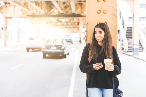 芝加哥一个站在繁忙道路旁笑着的多种族女孩 一个年轻女子的画像 目光从相机上移开 拿着一杯咖啡和智能手机 旁边的汽车在街上经过 — 图库照片