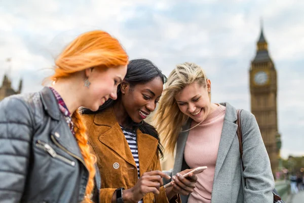 Multiracial group of girls in London looking at mobile phone with Big Ben on background. Three women in London having fun together. Travel and friendship concepts