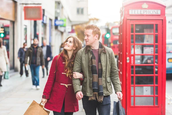 ロンドンでのハッピーカップルショッピングとウォーキング 忙しい通りの近くの赤い電話ボックスの隣に笑顔の若い女性と男 ライフスタイルの概念 — ストック写真