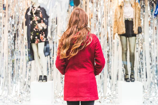 城市里穿着红色外套购物的年轻女人的后视镜 姜黄头发的年轻女人在商店橱窗前朝里看 生活方式的概念 — 图库照片