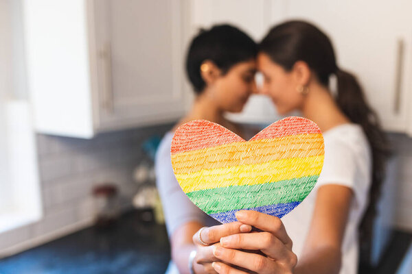 Authentic shot of happy lesbian couple holding a heart shaped rainbow flag and hugging - lesbian couple at home enjoying life together