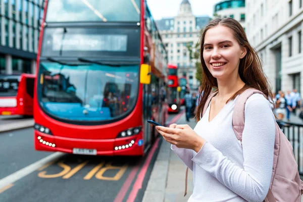 Smiling woman with smartphone at bus stop in London - Portrait of a smiling girl using her phone to check bus timetable on a day out in London - Lifestyle and transportation concepts