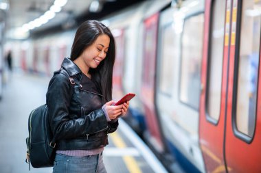 Yeraltındaki tren istasyonunda cep telefonu kullanan Çinli bir kadın treni bekleyen genç Asyalı bir kadın ve telefonda daktiloyla gülümseyen bir kadın.