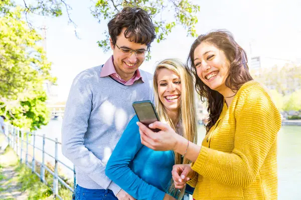 Three joyful friends enjoying social media content on a phone in a sunny park in Paris by Seine river and Eiffel tower - Friends laughing and sharing content on smartphone outdoors