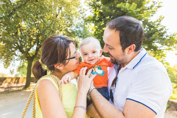 家族の外出中に愛情を持って赤ちゃんにキスハッピーな両親 公園で晴れた日を楽しむ楽しい家族 ロイヤリティフリーのストック写真