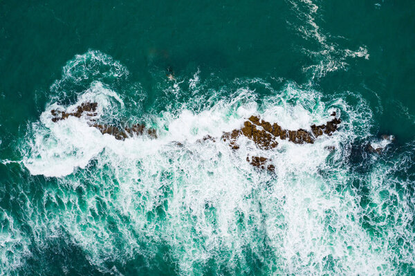 Top-down shot of turquoise ocean waves breaking against a rugged coast - Aerial view of crashing waves on rocky shoreline