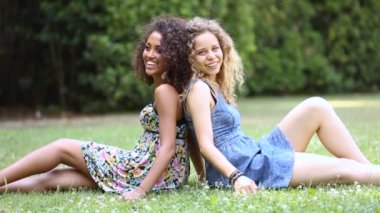 Parkta birlikte çok ırklı mutlu genç kadınlar - çimlerin üzerinde oturup gevşeyen sevimli kadın çift - melez siyah ve beyaz kıvırcık kızlar mutluluğu paylaşıyor