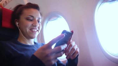 Uçakta akıllı telefonuyla müzik dinleyen bir kadın seyahat ve teknoloji kavramları, kulaklık takan genç bir kadın.