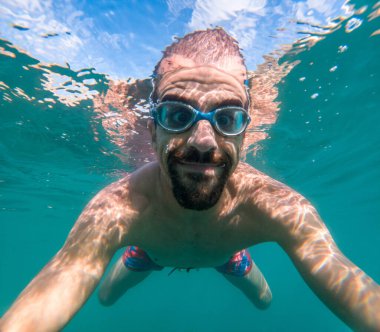 Yaz tatilinde deniz kenarında kristal berrak suyla yüzerken selfie çeken bir adam güneşli gün sporu aktivitesi.