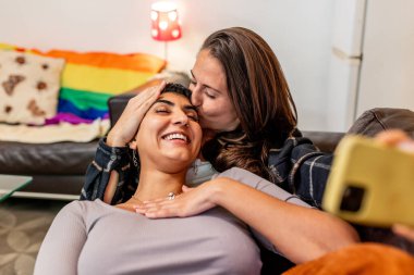 Birlikte eğlenen iki kadın. Kanepede selfie çekiyorum. Gökkuşağı bayrağıyla oturma odasında akıllı telefonlarıyla anı yakalarken sevgi ve mutluluk saçıyorlar.