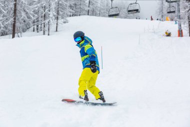 Snowboardcu kayak merkezindeki karlı bir yamaçtan kayıyor, tam teçhizatlı ve ustalıkla tahtayı manzaralı bir zemine doğru hareket ettiriyor. Kış sporlarının aktif yaşam tarzının tadını çıkarın.