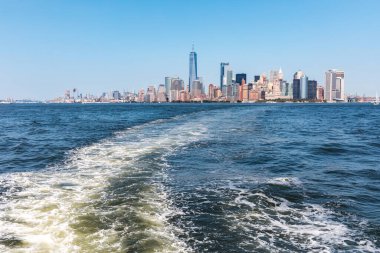 Feribot, güneşli bir yaz günü New York 'ta Manhattan ufuk çizgisine doğru köpüklü suda süzülür. Modern gökdelenlerin manzarası ve rıhtım boyunca tarihi işaretler sunuyor.