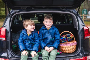 Mavi ceketli iki genç kardeş bir arabanın bagajında oturmuş piknik sepetinin yanında mutlu bir şekilde gülümsüyorlar. Sahne, çocukluk macerasını ve beraberliği bir yolculuk beklentisiyle yakalıyor.