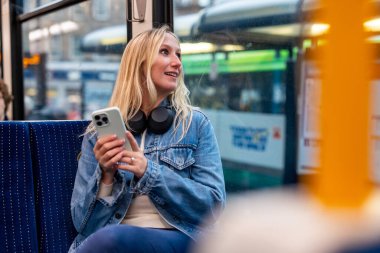 Kulaklıklı sarışın bir kadın akıllı telefon kullanırken müziğin keyfini çıkarıyor. Şehir içi seyahati sırasında otobüste rahatça oturuyor.