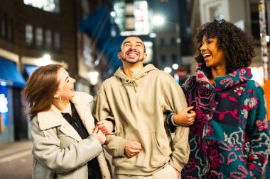 Three smiling diverse friends walking arm in arm, laughing together in an urban street at night, enjoying friendship and togetherness