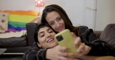 Mutlu lezbiyen bir çiftin yeni evlerindeki koltukta selfie çekerken ve homoseksüel evli eşcinsel çiftin arka planda gökkuşağı gururu bayraklarıyla eğlenirken sahnelenen otantik sinematik çekimleri.