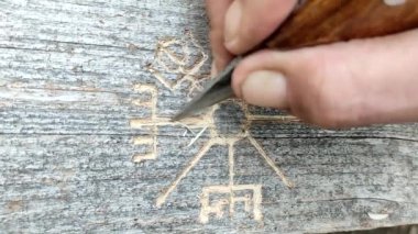 A mans hand with a knife carves the Viking rune of the Vegvisir compass on hard wood, close-up