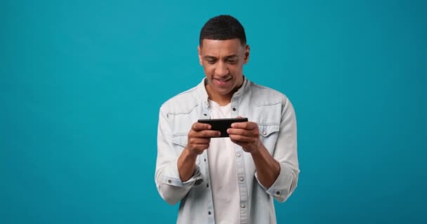 オンラインゲーム 青のスタジオの背景に立ってスマートフォンでモバイルビデオゲームをプレイする若いアフリカ系アメリカ人のゲーマー Dci 二重スローモーション — ストック動画