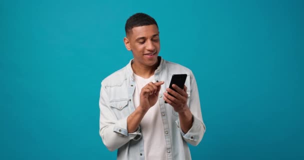 若い笑顔青いスタジオの背景に立って携帯電話を使用してアフリカ系アメリカ人の男 Dci 二重スローモーション — ストック動画