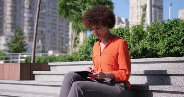 幸せな若いアフリカ系アメリカ人女性が屋外に座って新しいアイデアに触発インターネットをサーフィンデジタルタブレットパッドを使用して — ストック動画