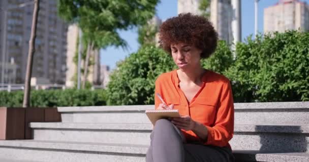 かわいい魅力的な女性アーティストはノートにノートを書くか何かを描く外の石の通りの階段に座っている — ストック動画