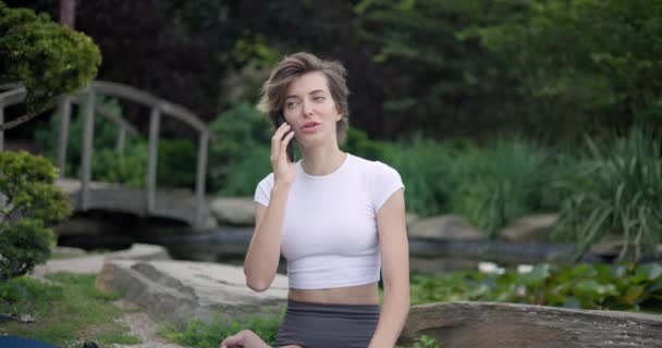 若い美しい女性は マットの上に座っている屋外でヨガ演習をしている間 電話で話しています — ストック動画