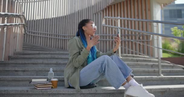 若いアフリカ系アメリカ人女性学生は ステップ上の屋外に座っている間 携帯電話でビデオ通話をする — ストック動画