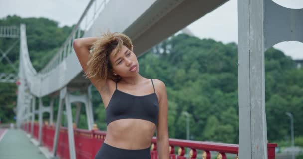 若いアフリカ系アメリカ人の女性に合う暖まると橋の上をジョギングする前にストレッチ — ストック動画