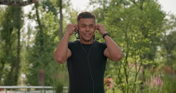 若いスポーティなアフリカ系アメリカ人のスポーティな男は 市立公園で屋外でジョギングする前にイヤホンを置きます スローモーション — ストック動画