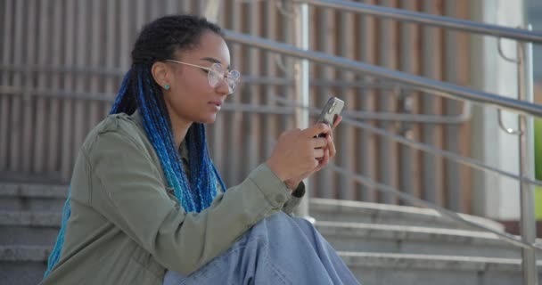 若いアフリカ系アメリカ人女性の学生は ステップ上の屋外に座っている間 携帯電話にメッセージを書くタイプ — ストック動画
