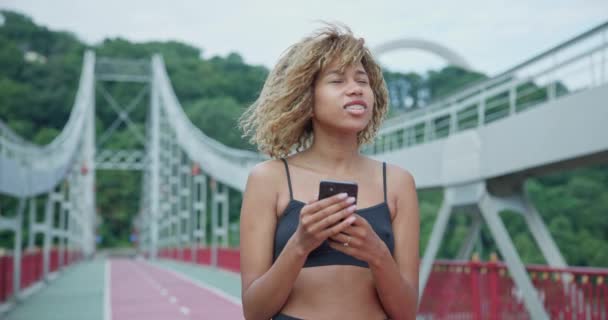 若いアフリカ系アメリカ人女性はヘッドフォンを着て 街の橋で屋外でジョギングする前に電話で音楽を選択します — ストック動画