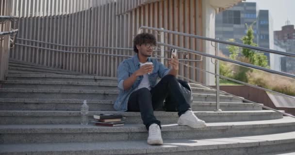 若いインド人の学生が階段に座り 屋外でコーヒーを飲み 電話で話す スローモーション — ストック動画