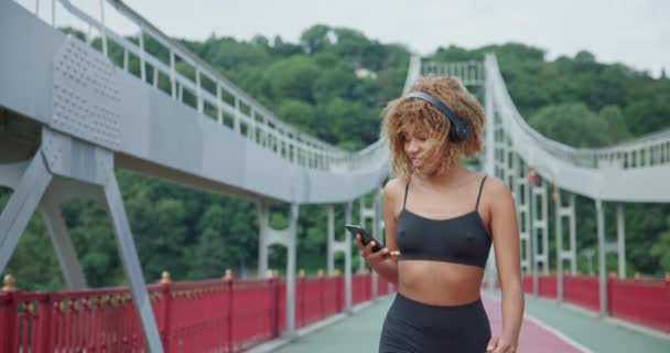 若いアフリカ系アメリカ人女性が街の橋でジョギングアウトドアでヘッドフォンを着用 — ストック動画