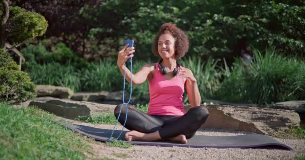 若いアフリカ系アメリカ人女性が屋外でヨガを練習するマットに座ってビデオ通話をする — ストック動画