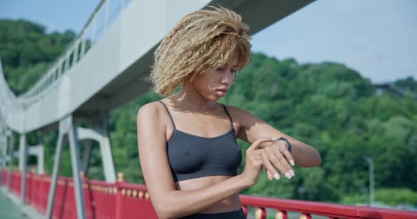 スポーティなアフリカ系アメリカ人女性が彼女のスポーツブレスレットを見て 屋外でジョギングした後 フィットネストラッカーのデータをチェック — ストック動画
