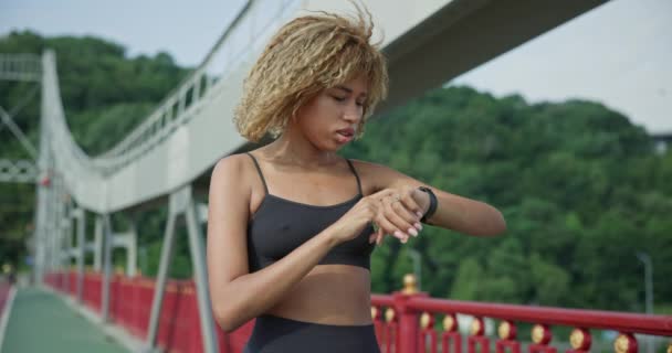 スポーティなアフリカ系アメリカ人女性が彼女のスポーツブレスレットを見て 屋外でジョギングした後 フィットネストラッカーのデータをチェック — ストック動画
