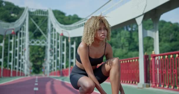 若いアフリカ系アメリカ人女性は ジョギングのために準備する都市の橋の上に座ってスニーカーをレース — ストック動画
