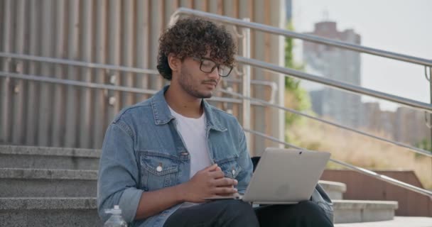 若いハンサムなインドのヒンドゥー教徒の学生フリーランサーは ラップトップコンピュータを使用してビデオ通話をするステップで屋外に座っています スローモーション — ストック動画