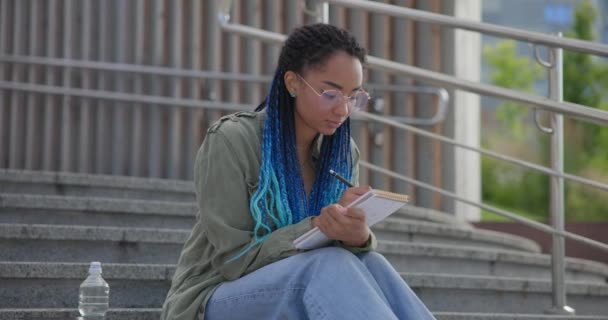 若いアフリカ系アメリカ人の女子学生が屋外の階段に座ってノートをノートに書きながら — ストック動画
