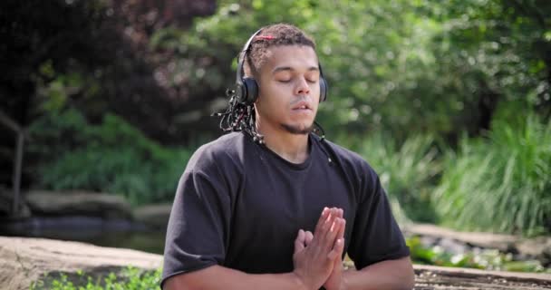 若いアフリカ系アメリカ人のスピリチュアリティマンは 公園の屋外で座って音楽を聴いているヨガアサナ瞑想 — ストック動画
