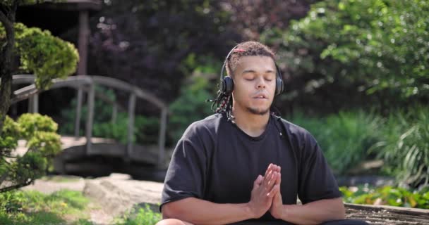 若いアフリカ系アメリカ人のスピリチュアリティマンは 公園の屋外で座って音楽を聴いているヨガアサナ瞑想 — ストック動画