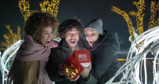 Возбужденные Счастливые Друзья Открывает Подарочную Коробку Луком Стоя Гирляндах Огни — стоковое видео