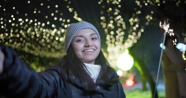 ライトとガーランドの背景に立っているスパークラーを保持するビデオコールを作る若いインドの女性 新年とクリスマスのコンセプト — ストック動画