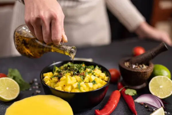 マンゴーサルサを調理 黒セラミックボウルでスライスした食材にオリーブオイルを注ぐ女性シェフ — ストック写真