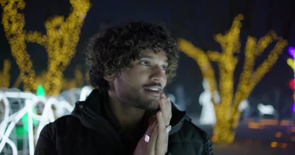 ガーランドやライトの背景に冬の夜に屋外に立っている若いインド人男性 — ストック動画