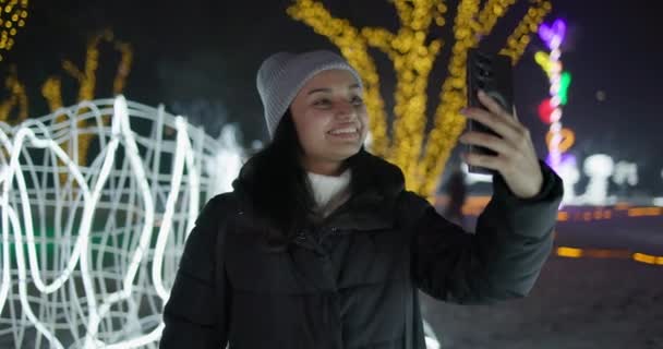 美しいインドの作るビデオ通話ガーランドやライトの背景の冬の夜に屋外に立っている電話 — ストック動画