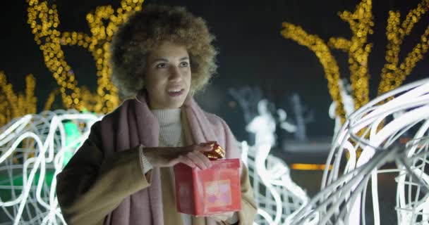若いアフリカ系アメリカ人女性は ギフトボックスを開き ガーランドに立って屋外の背景を明るくします クリスマス休暇 ロイヤリティフリーのストック動画